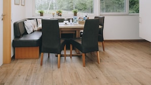 A cozy room with a wooden floor, a black sofa, a dining table with chairs, and a window with a view of greenery.