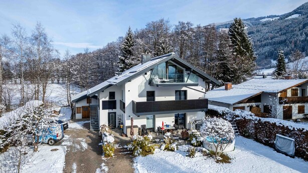 A modern two-story house with a large glass balcony, surrounded by snow-covered trees and a mountainous backdrop.