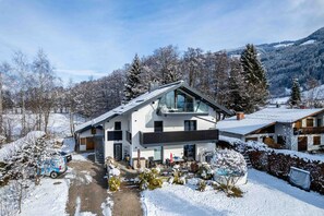 Une maison moderne à deux étages, dotée d’un grand balcon en verre, entourée d’arbres couverts de neige et surplombant un paysage montagneux.