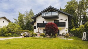 A modern two-story house with a balcony, surrounded by lush greenery and a well-maintained lawn.