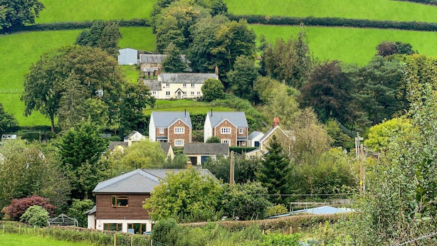 A rural landscape with rolling green hills, a large house with multiple buildings, and surrounding trees.