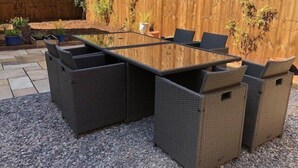 A rectangular glass-top table with dark wicker chairs, set on gravel.