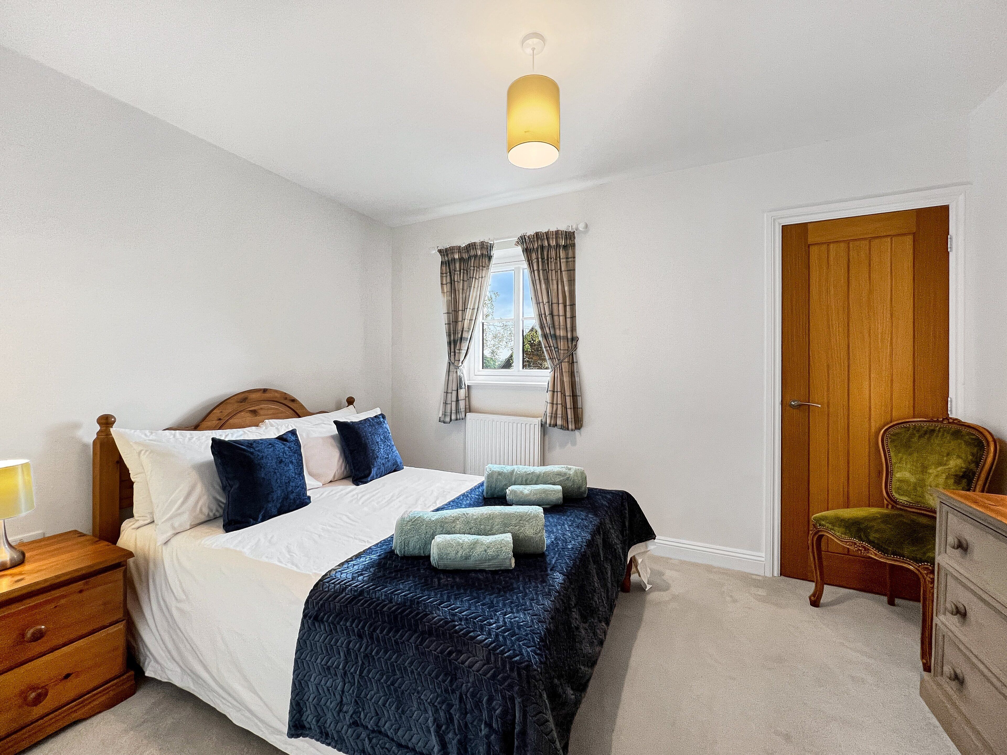 Een slaapkamer met een houten bed, nachtkastje, dressoir, raam met gordijnen en een deur.