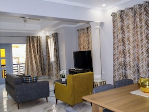 Relax in this stylish living room with a sofa, armchairs, and a dining table, featuring a flat-screen TV and a potted plant.