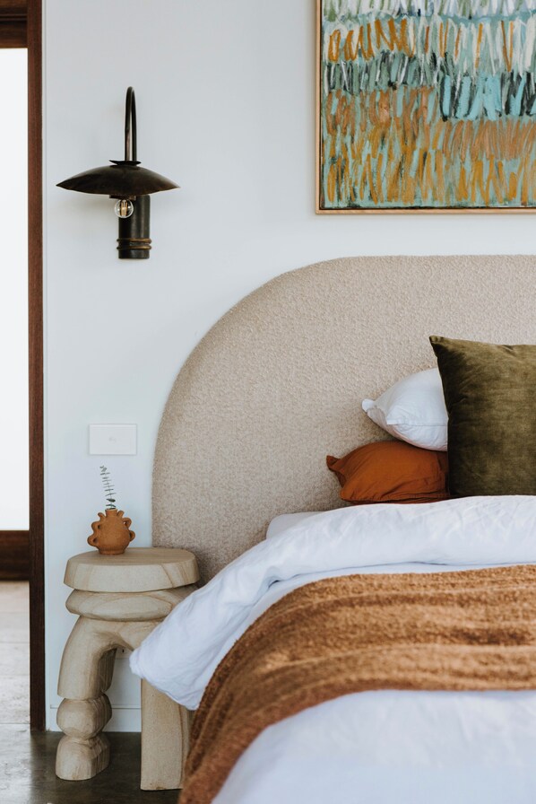 Een slaapkamer met een groot bed, een nachtkastje met lamp, een decoratieve kruk en een wanddecoratie.