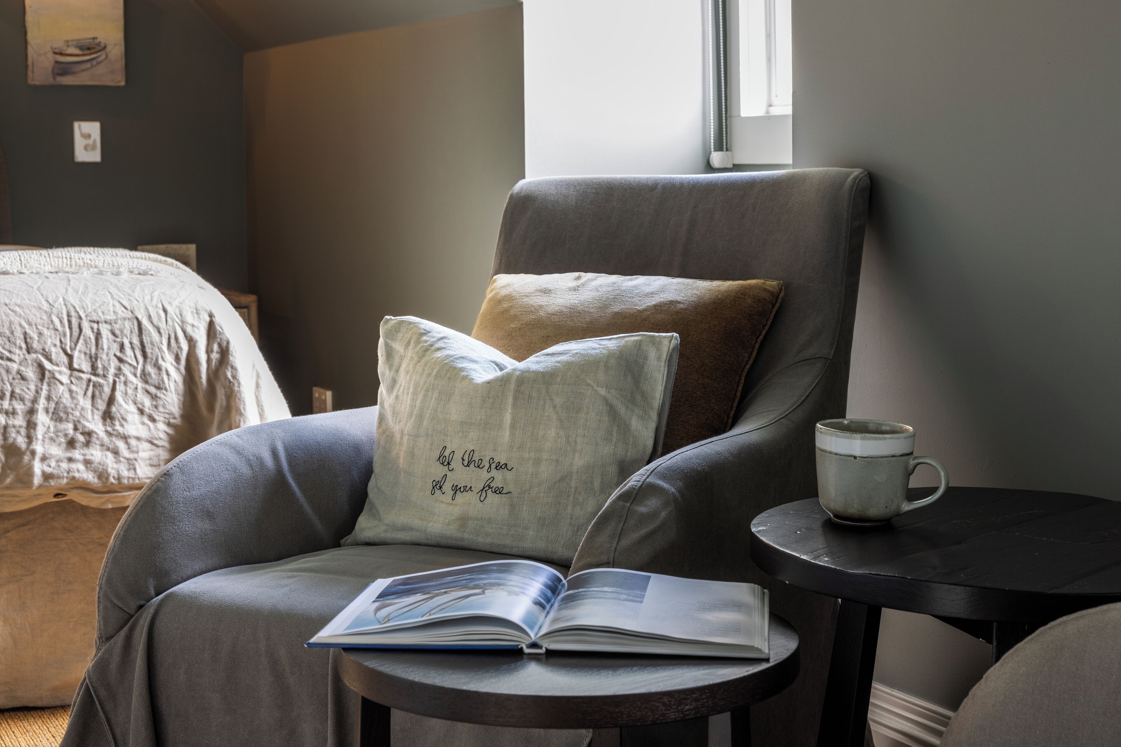A cozy room with a grey armchair, a small round table with a book and a mug, and a bed with a beige comforter.