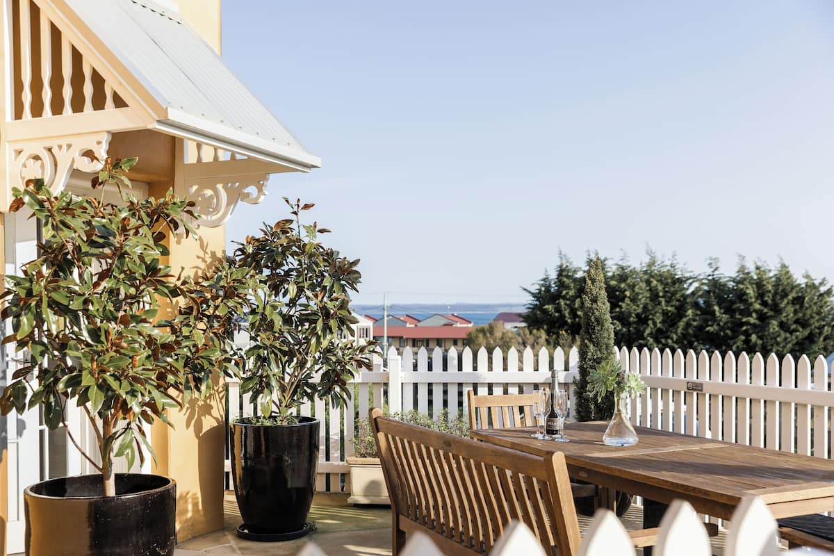 An outdoor patio with a wooden table and chairs, potted plants, and a white picket fence.