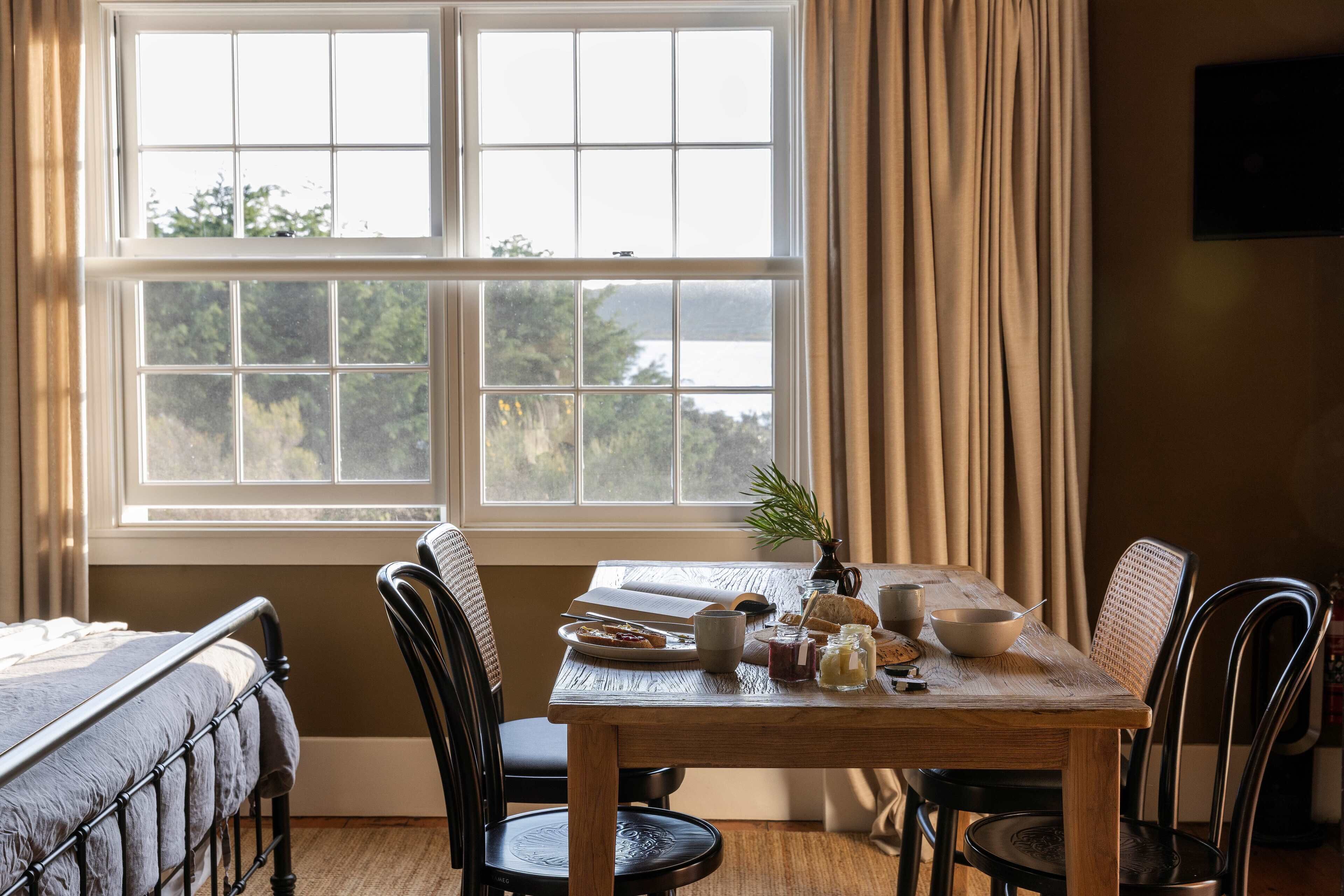 A dining area with a table set for two, a window with curtains, and a bed in the background.