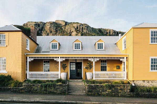 Um edifício amarelo com varanda branca, degraus de pedra e montanhas ao fundo.