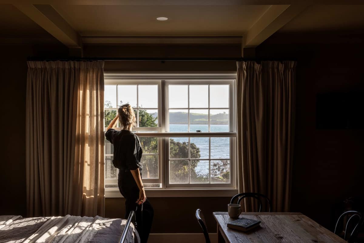 A room with a large window, curtains, a table with a chair, and a person standing by the window.