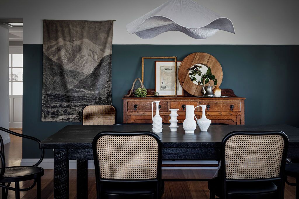 Un comedor con una mesa de madera oscura, dos sillas de mimbre, un aparador de madera con adornos y un tapiz grande en la pared.