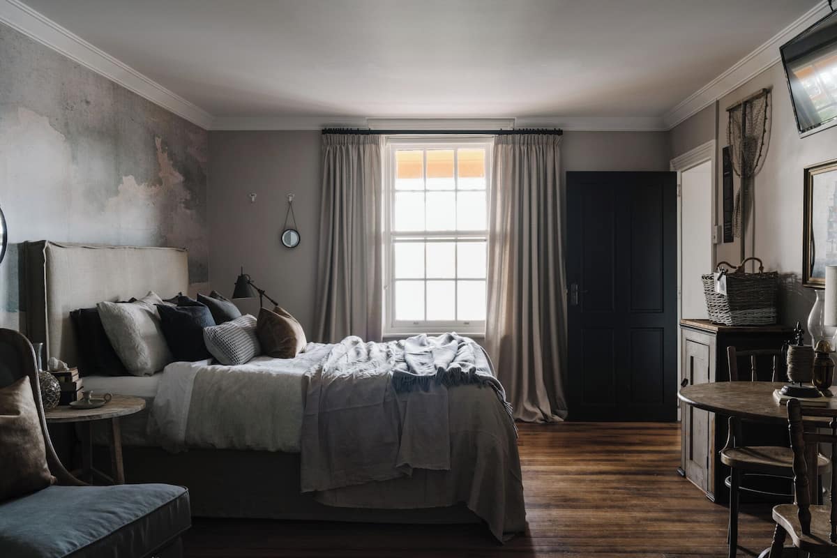 A bedroom with a bed, bedside table, lamp, and a view of the outside through a window.