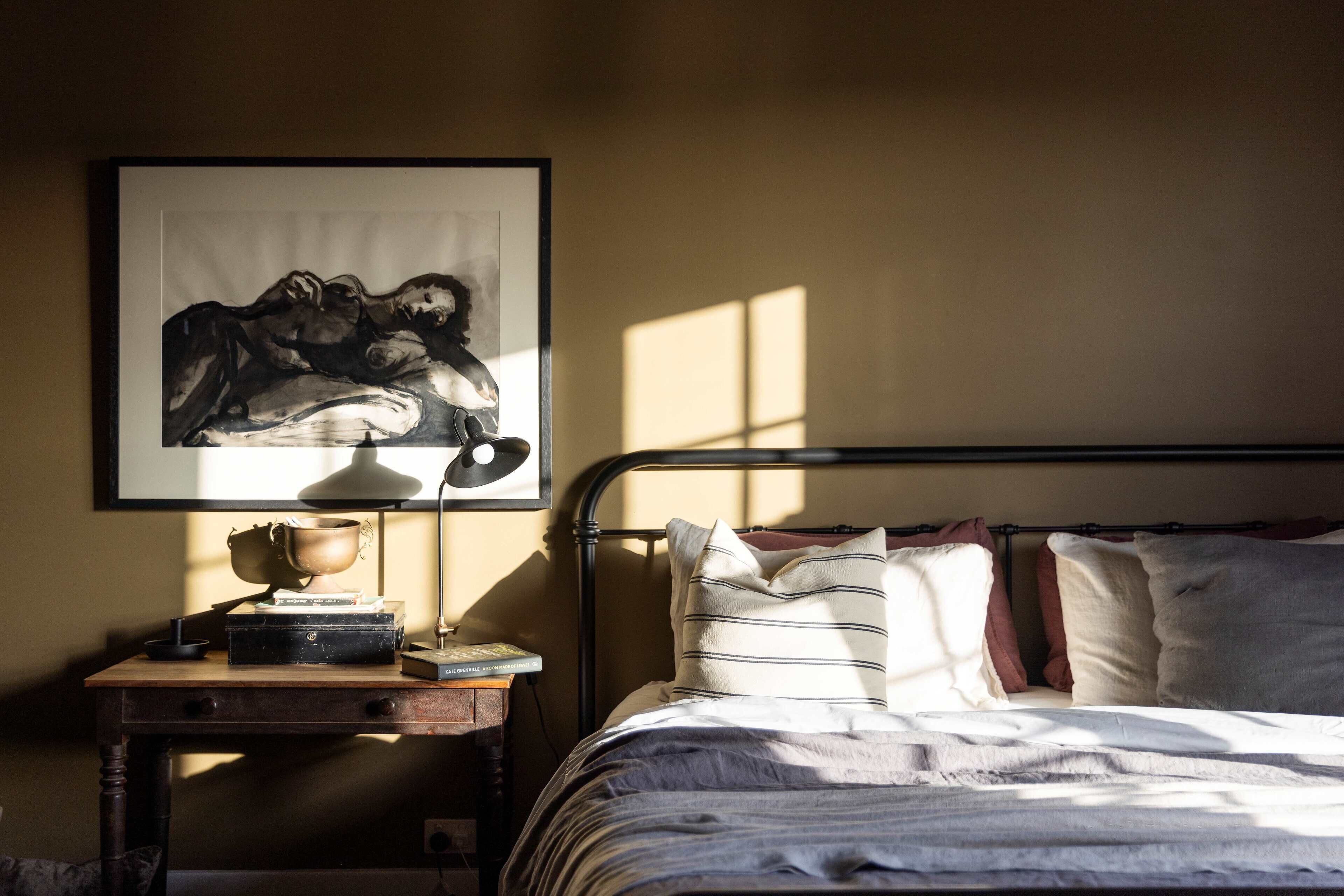 Un dormitorio con cama, almohadas, una mesita de noche con lámpara, una obra de arte enmarcada y una ventana que deja entrar luz.
