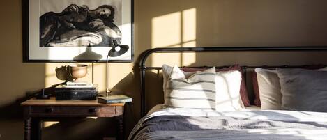 A bedroom with a bed, pillows, a bedside table with a lamp, a framed artwork, and a window casting light.