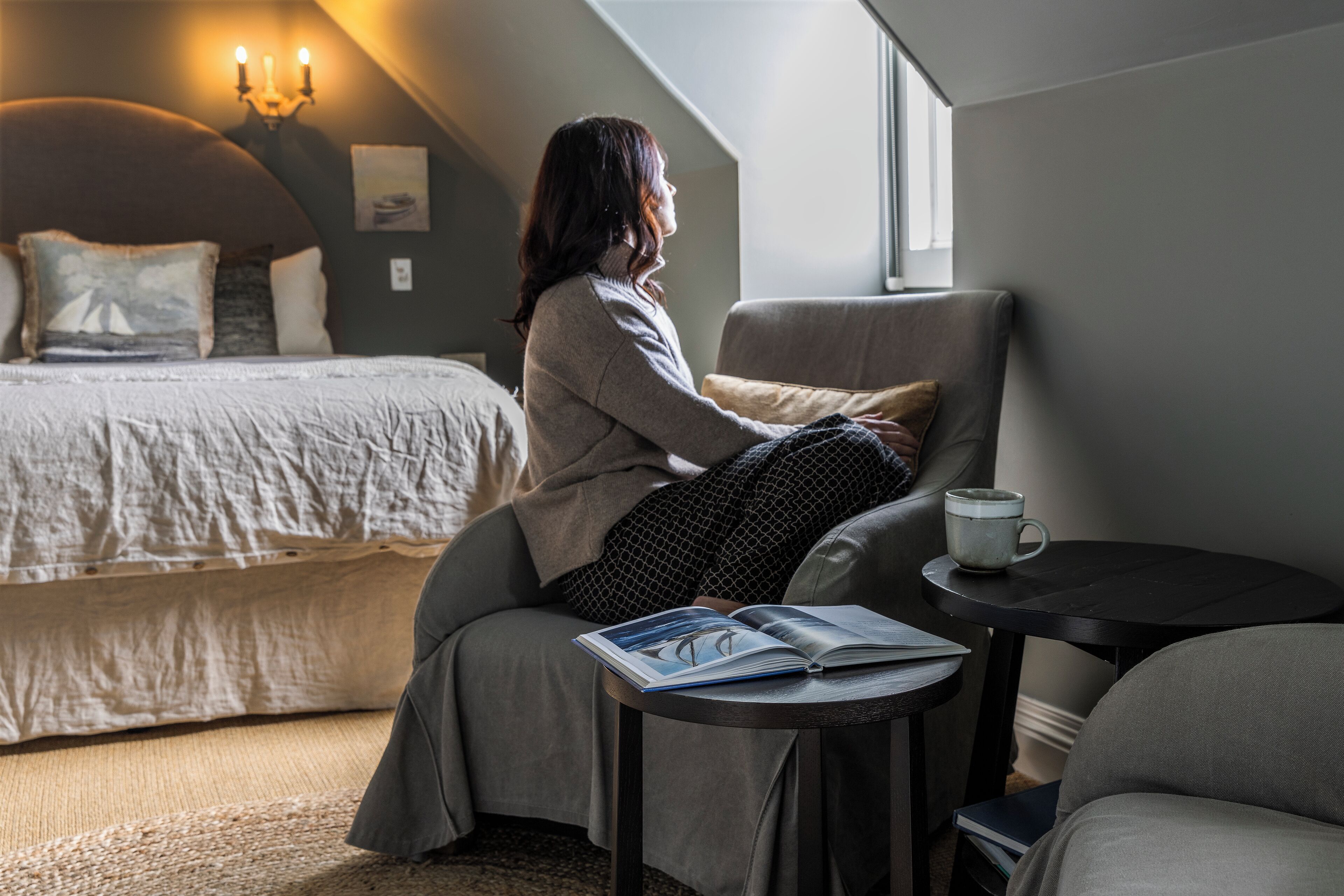 Una habitación acogedora con una cama, un sillón de lectura, una mesita con un libro y una taza, y una ventana que deja entrar luz natural.
