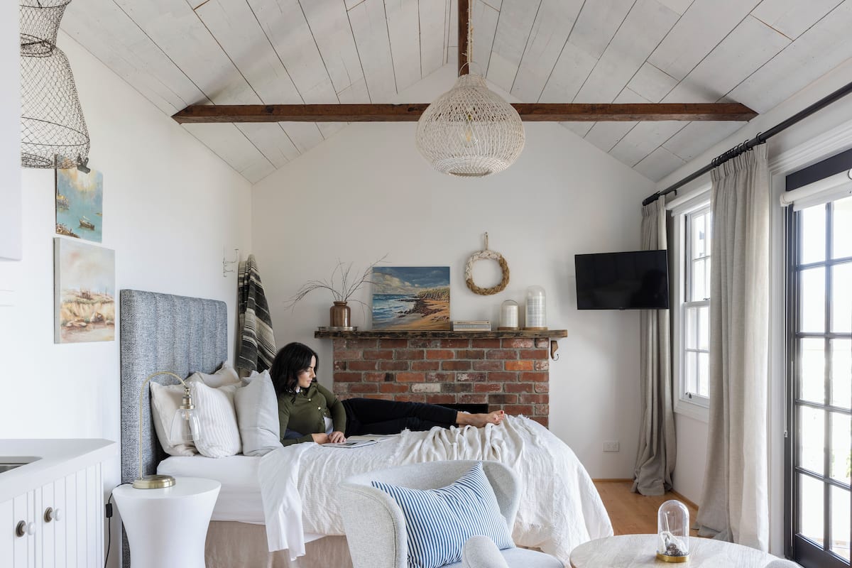 A cozy bedroom with a brick fireplace, a bed with a white headboard, a white dresser, and a large window with curtains.