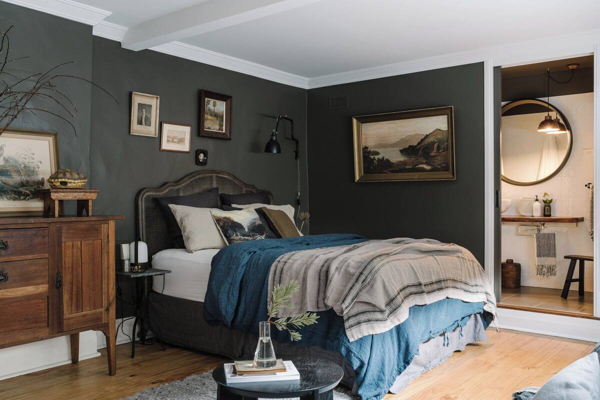 A bedroom with a dark wall, a wooden dresser, a bed with a blue blanket, and a large mirror.