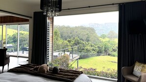 A bedroom with a large bed, a view of a green landscape, a balcony with a railing, and a chair with a cushion. - Grand View Luxury Villa (Kangaroo Valley)