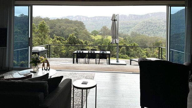 A modern living room with a large sliding glass door leading to a deck with a view of a forested mountain range. - Grand View Luxury Villa (Kangaroo Valley)