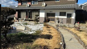 Une maison traditionnelle japonaise, avec un toit recouvert de tuiles, un jardin agrémenté d’un chemin de pierres et un petit espace extérieur aménagé pour s’asseoir.