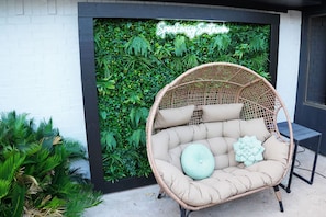 Una silla de mimbre con cojines frente a una pared cubierta de follaje verde y un letrero de neón.