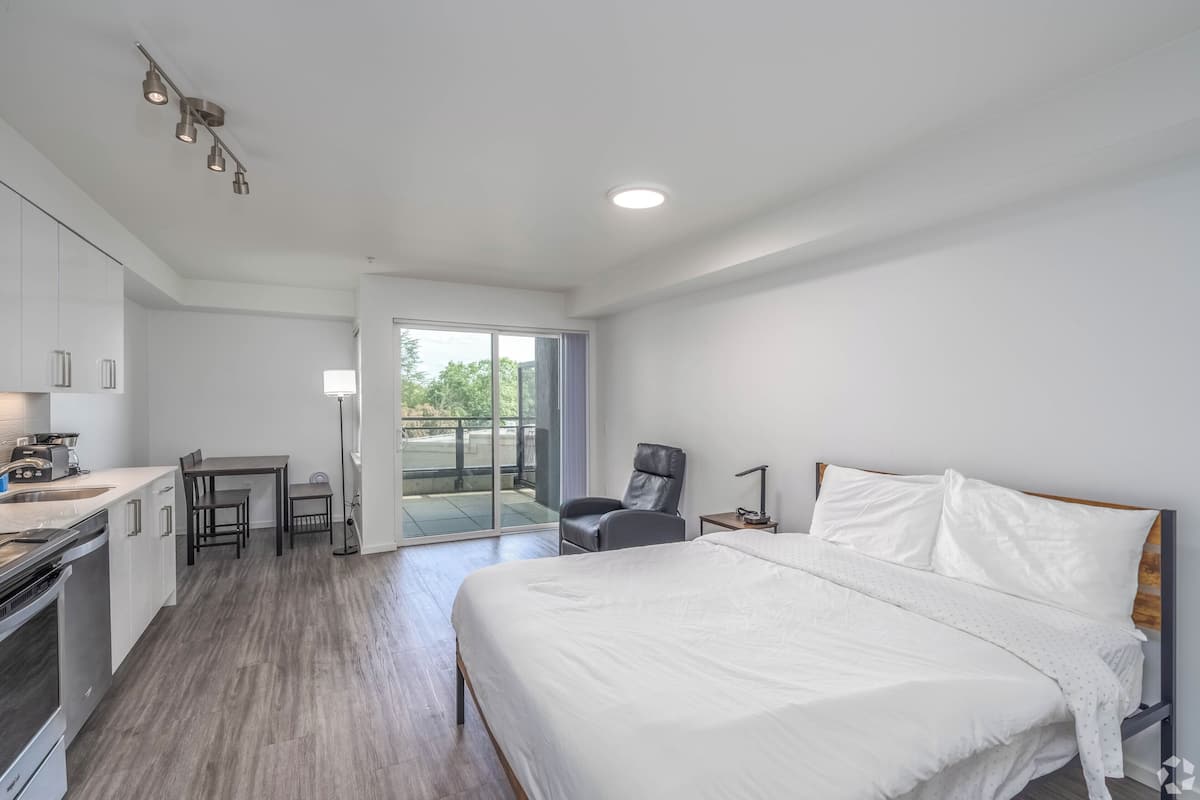 A modern hotel room with a large bed, a small kitchenette, a dining area, and a sliding glass door to a balcony.