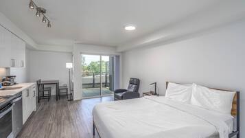 A modern hotel room with a large bed, a small kitchenette, a dining area, and a sliding glass door to a balcony.