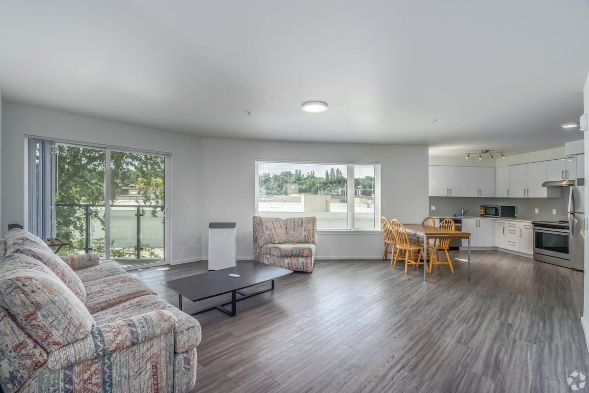 A modern living room with a sofa, a coffee table, and a dining area with chairs and a table. There is a kitchen with cabinets and appliances. Large windows provide natural light and a view of greenery outside.