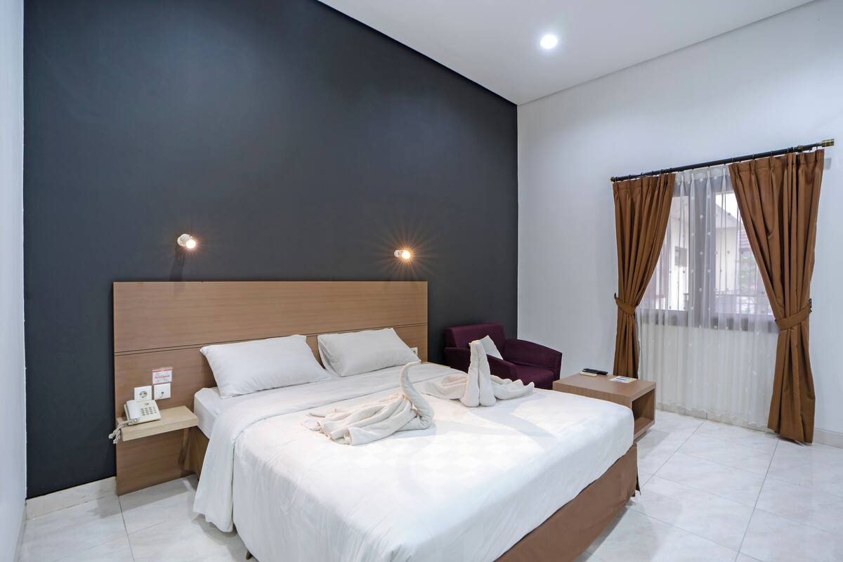 A modern hotel room with a large bed, a bedside table with a phone, a chair, and a window with curtains.
