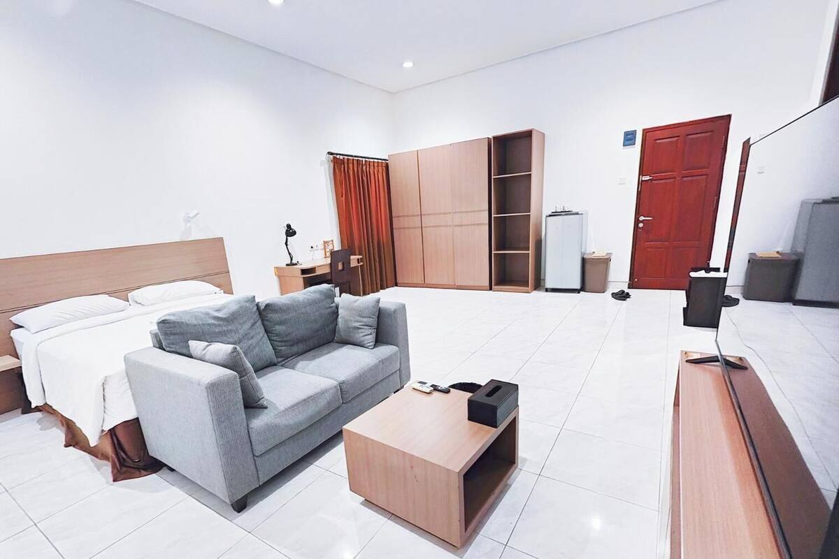 A modern hotel room with a bed, grey sofa, wooden coffee table, desk, wardrobe, and a TV stand.