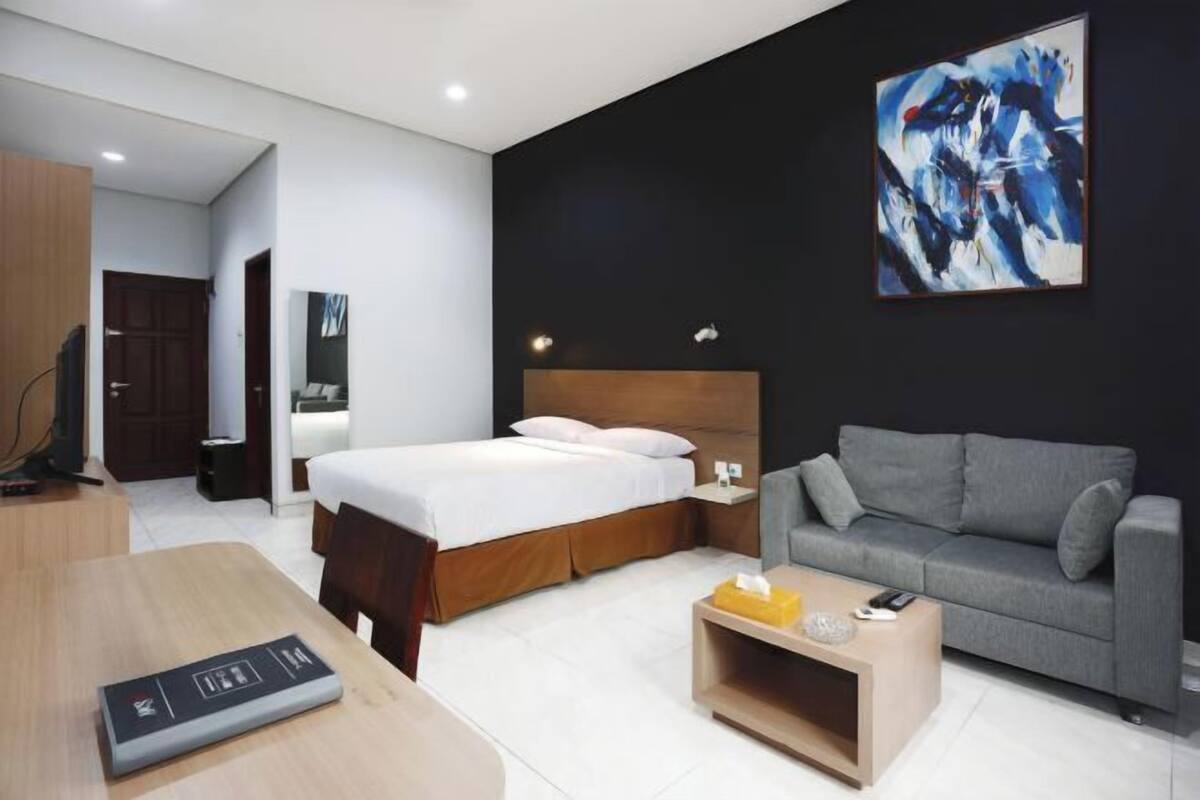A modern hotel room with a bed, a sofa, a desk, and a painting on the wall.