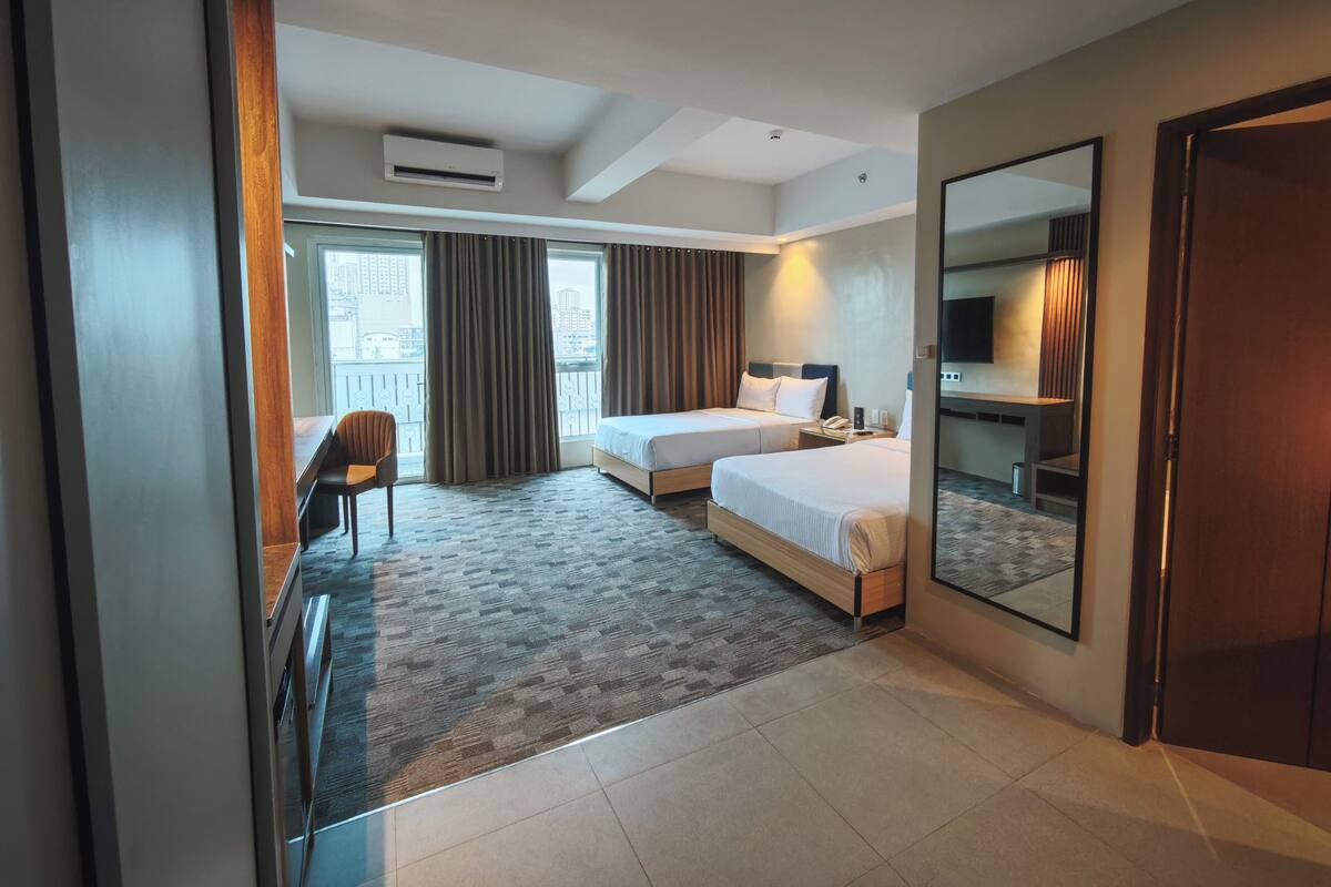 A hotel room with two beds, a desk, a chair, a large mirror, and a view of the city through the window.
