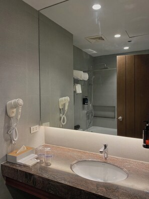 A modern bathroom with a granite countertop, a sink, a mirror, a hairdryer, a towel rack, and a shower. - The Tribute Hotel (Quezon City)