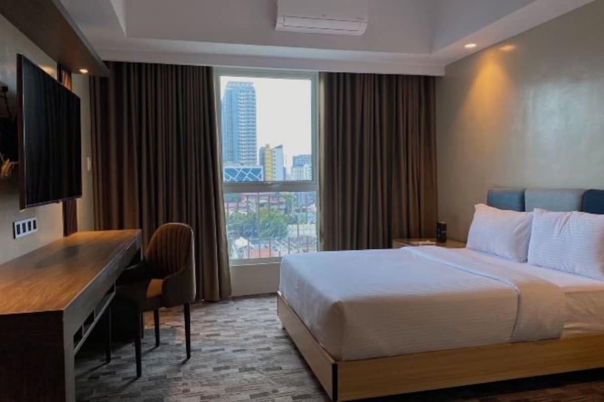 A hotel room with a bed, a desk, a chair, a TV, and a window with curtains.