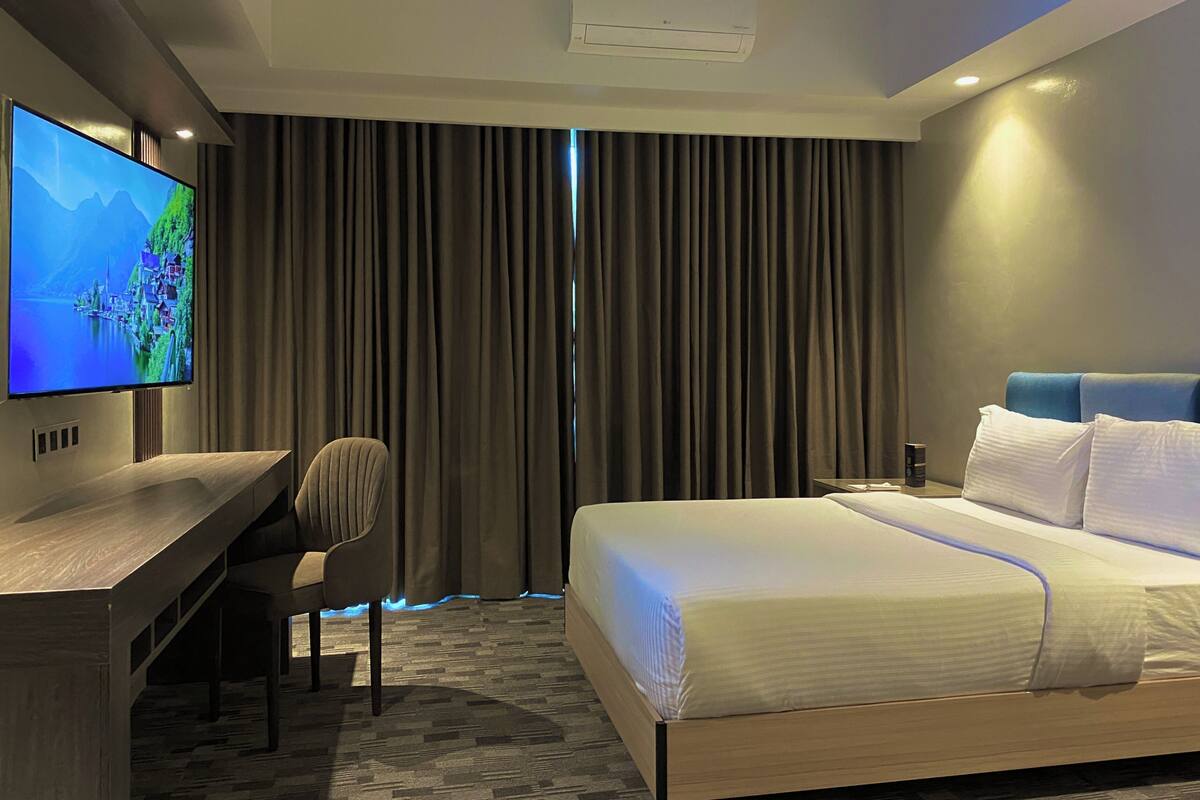 A hotel room with a bed, a desk with a TV, a chair, and a window with curtains.