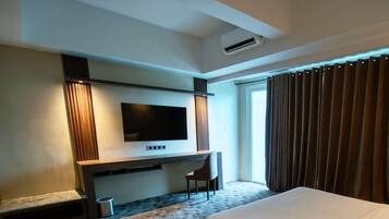 Quarto de hotel moderno com uma cama grande, televisão de tela plana na parede, uma escrivaninha pequena com cadeira e uma janela com cortinas.