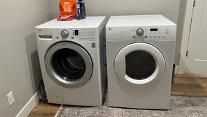 Deux machines à laver à chargement frontal se trouvent dans une buanderie, sur un tapis gris, avec une porte blanche en arrière-plan.