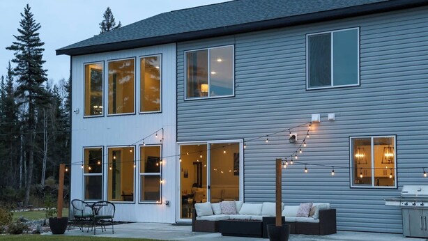 Una casa moderna de dos pisos con ventanas amplias, un patio con mobiliario exterior y luces colgantes.