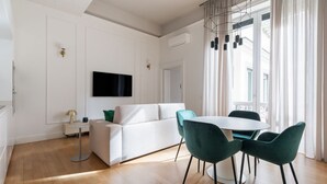 A modern living room with a white sofa, a round dining table with green chairs, a flat-screen TV mounted on the wall, and a ceiling light fixture. - Vittorio Emanuele | Luxe Flat (Roma)
