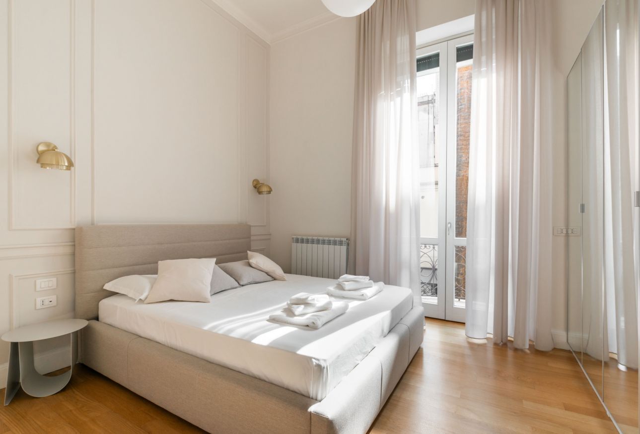 A modern bedroom with a large bed, a small bedside table, a radiator, and a view of the outside through a glass door.
