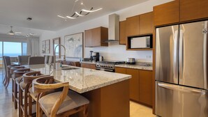 Une cuisine moderne dotée d’un îlot central, d’appareils électroménagers en acier inoxydable, de chaises en bois et d’une grande fenêtre donnant sur l’océan.
