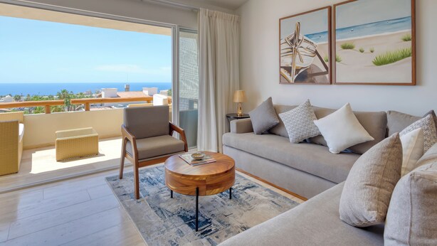 Un salon moderne avec un canapé, un fauteuil et une table basse, un balcon offrant une vue sur la mer, et deux œuvres d’art encadrées accrochées au mur.