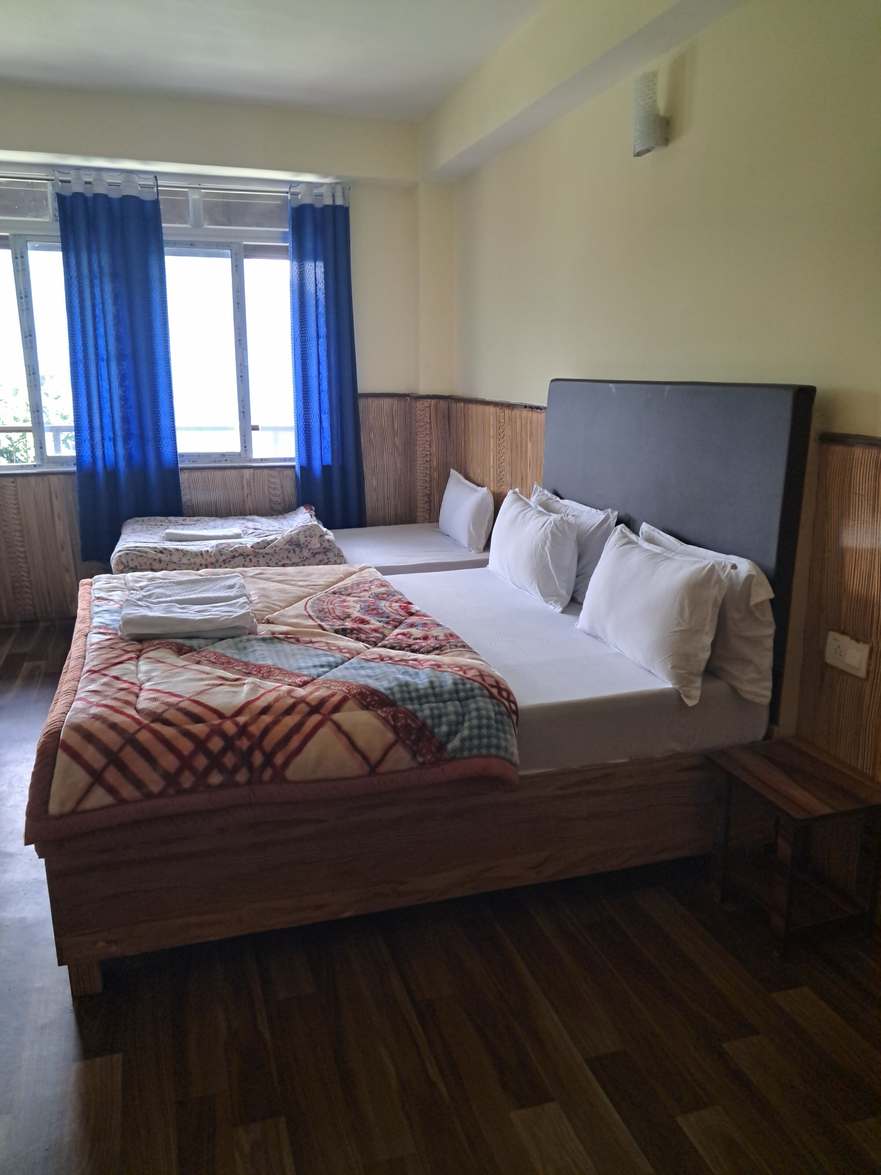 A hotel room with a large bed, two smaller beds, wooden flooring, a window with blue curtains, and a plain wall.