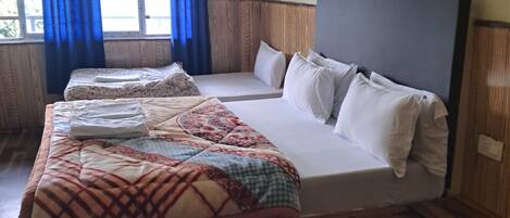 A hotel room with a large bed, two smaller beds, wooden flooring, a window with blue curtains, and a plain wall.