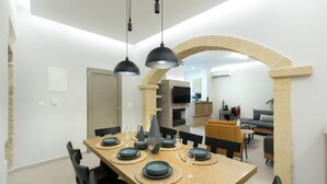 Un espace repas avec une longue table dressée pour six personnes, des chaises noires et une arche en pierre menant à un salon équipé d’un canapé et d’un téléviseur.