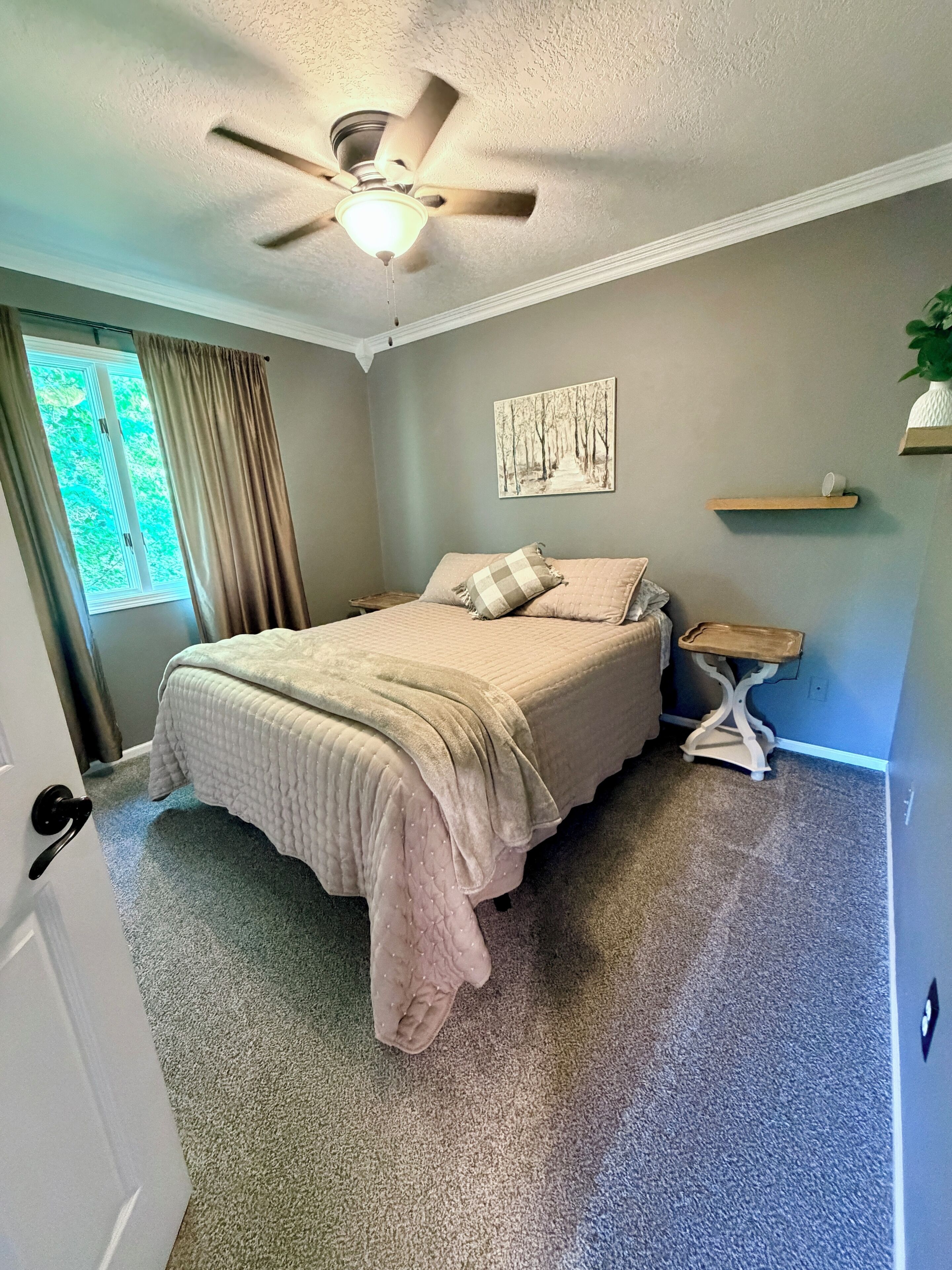 A bedroom with a bed, a ceiling fan, a window with curtains, a painting on the wall, and a small shelf.