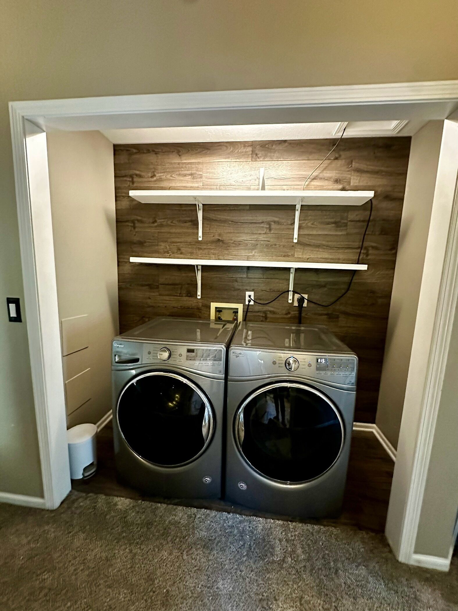 A laundry room with two front-loading washing machines, wooden shelves above, and a carpeted floor.