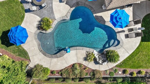 An aerial view of a residential swimming pool with a unique shape, surrounded by a patio area with outdoor furniture and umbrellas.
