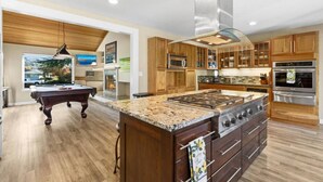 A modern kitchen with wooden cabinets, stainless steel appliances, a central island, and a pool table in the adjacent room.