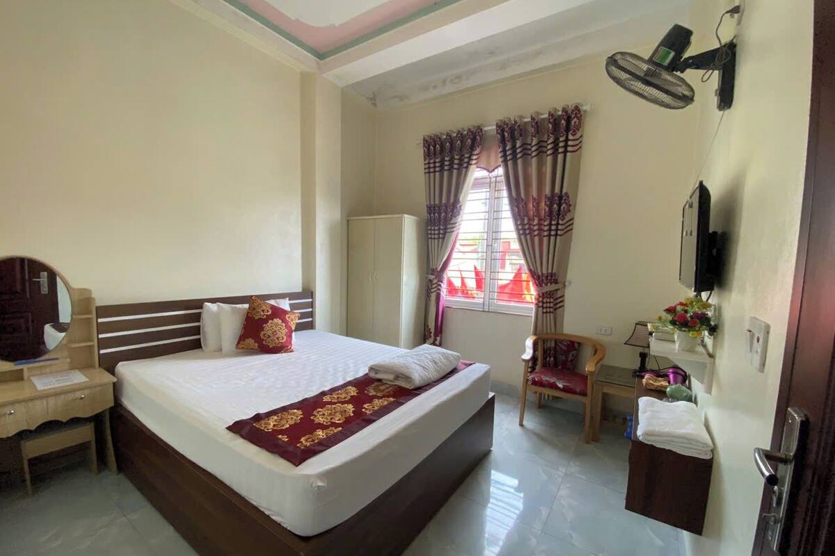 A hotel room with a large bed, a desk with a mirror, a chair, a wardrobe, and a small table with a lamp and flowers.